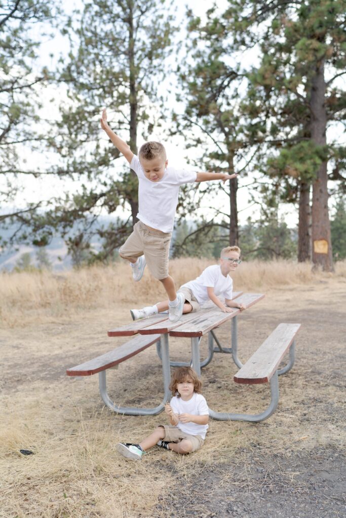 Three young boys in white tshirts and khaki shorts play on an outdoor table and chair arrangement.