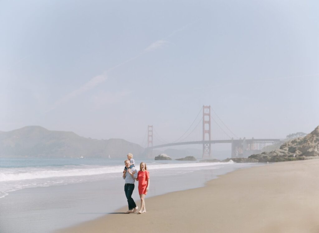 A married couple and their child at the Baker Beach with the Golden Gate Bridge in the background.
