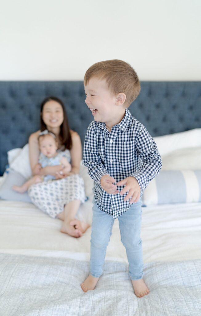 Little baby boy stands on his bed and smiles while his mom holds his newborn baby sister on her lap.