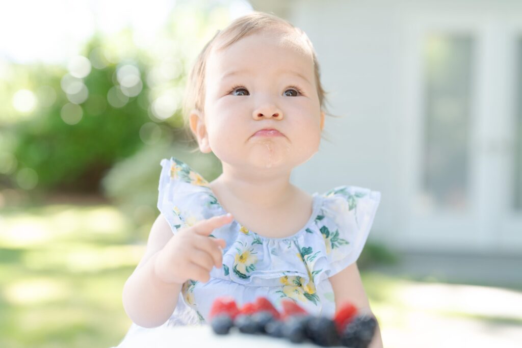 Baby daughter in a cute dress sits behind a white cake with straberries and black berries.