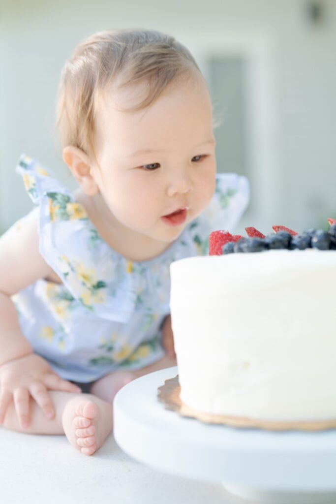 Adorable baby girl looks curiously at a small white cake, which seems gigantic besides her.