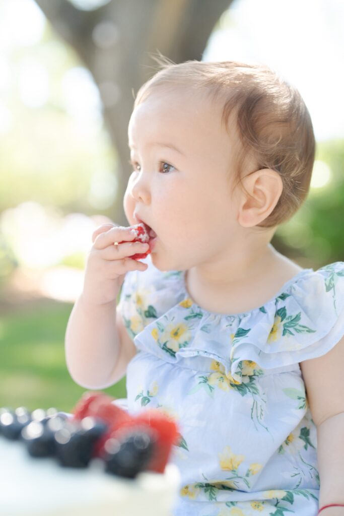 Baby sits behind a white cake and eats a strawberry.