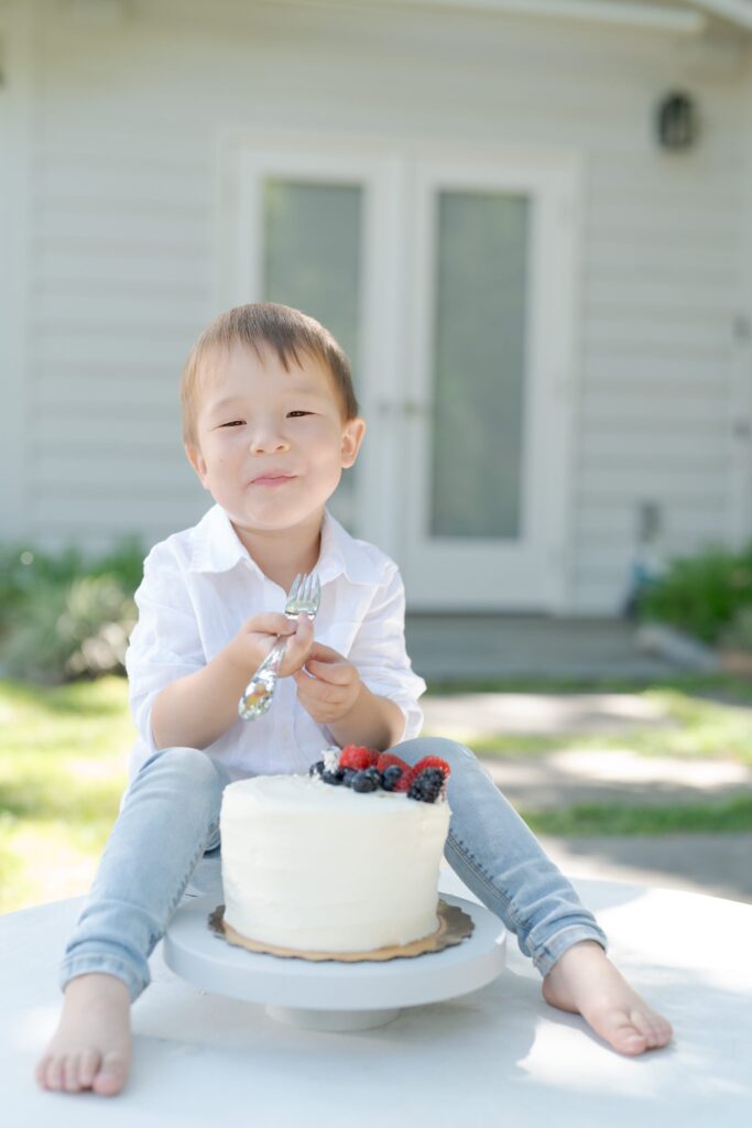 Baby boy holds a fork and sits behind a delicious-looking white cake.