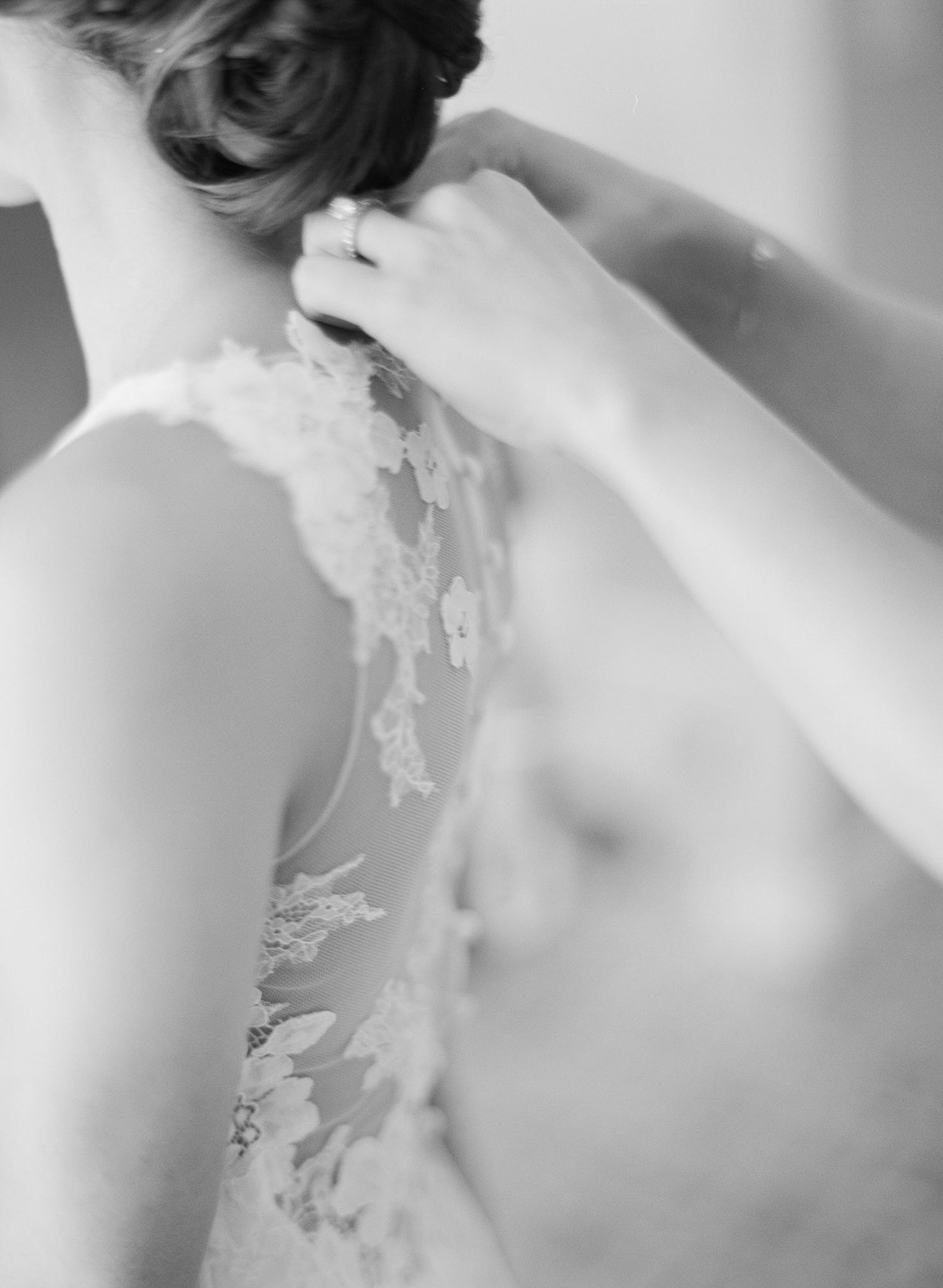 A black and white image of person buttoning a woman into her wedding dress.