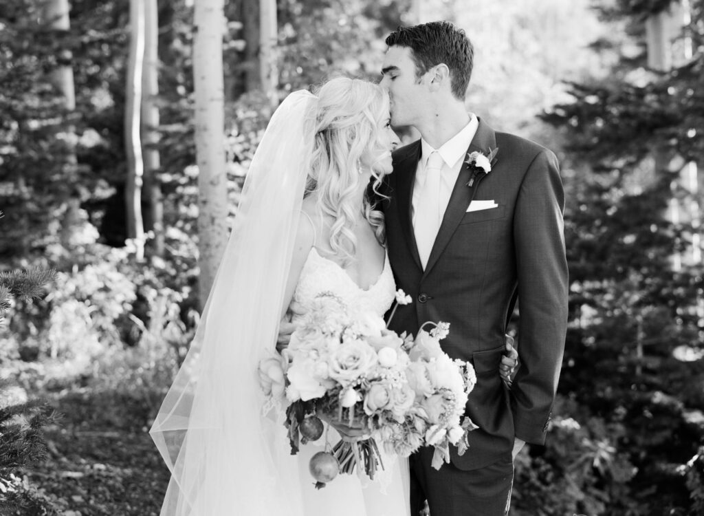 Grayscale picture of a bridegroom kissing his bride in bridal dress and a bouquet.
