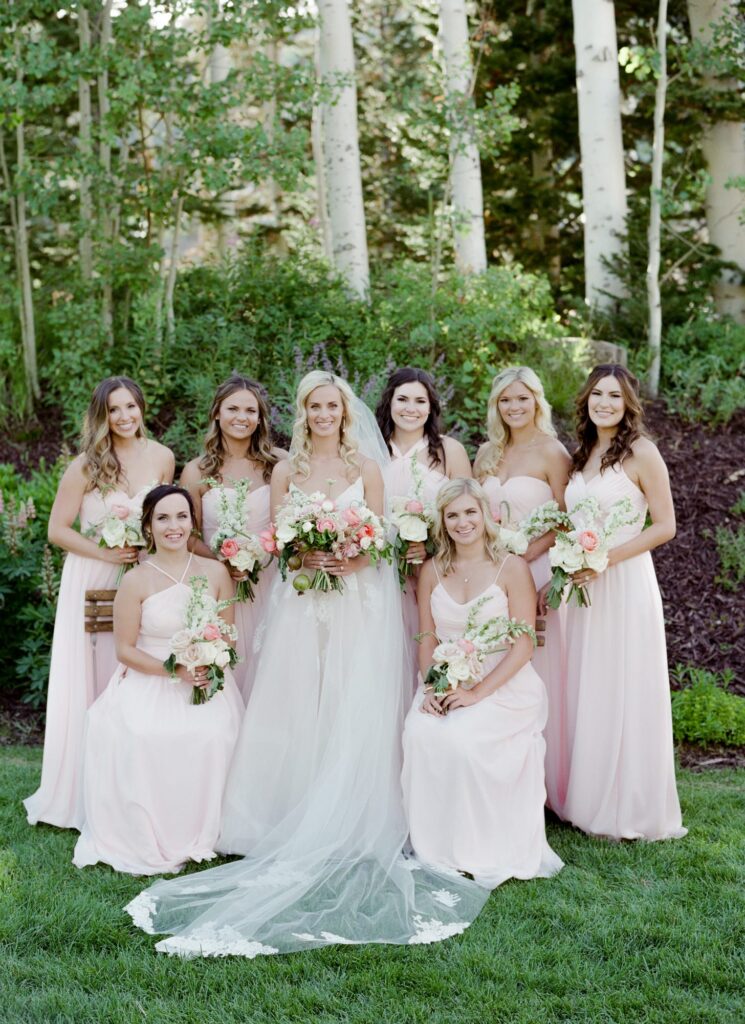 Bride poses for the camera outdoor among the woods with her bridesmaids holding flower bouquets.