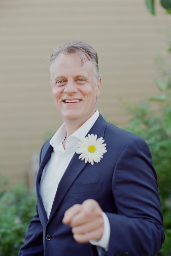 Groom with a daisy flower on his suit points at the camera.