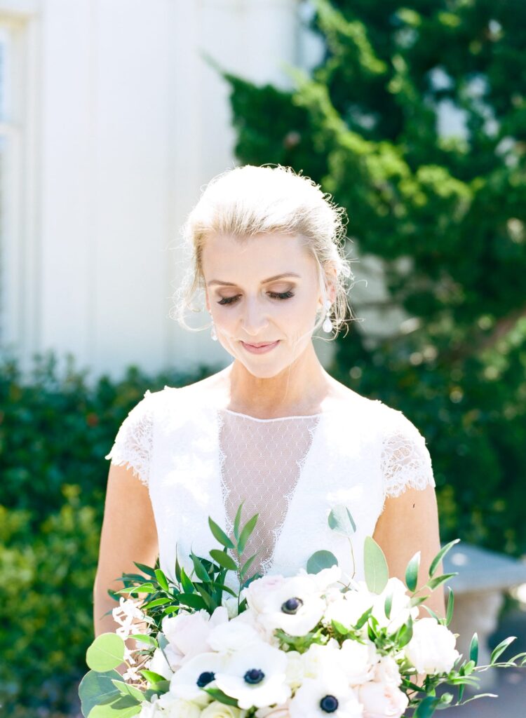 Bride in wedding gown looks down while holding a white flower bouquet.