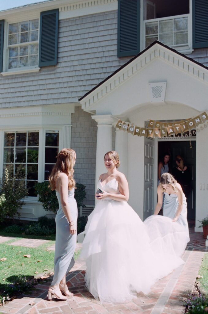 Bride comes outdoors in her wedding gown while a bridesmaid holds her train.