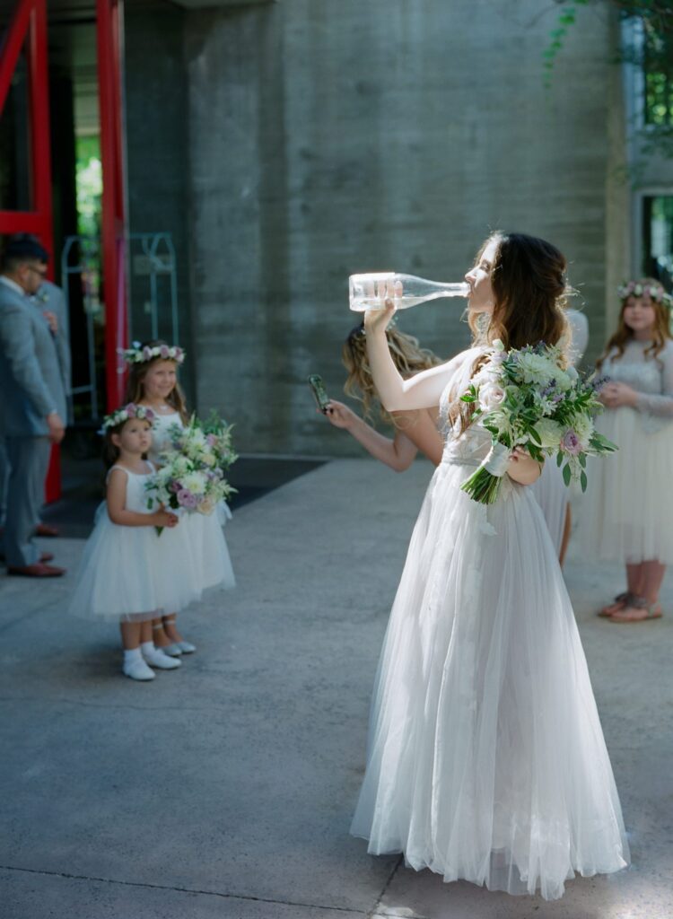 Bride in wedding dress drinks out of a glass bottle while little girls watch in the background.