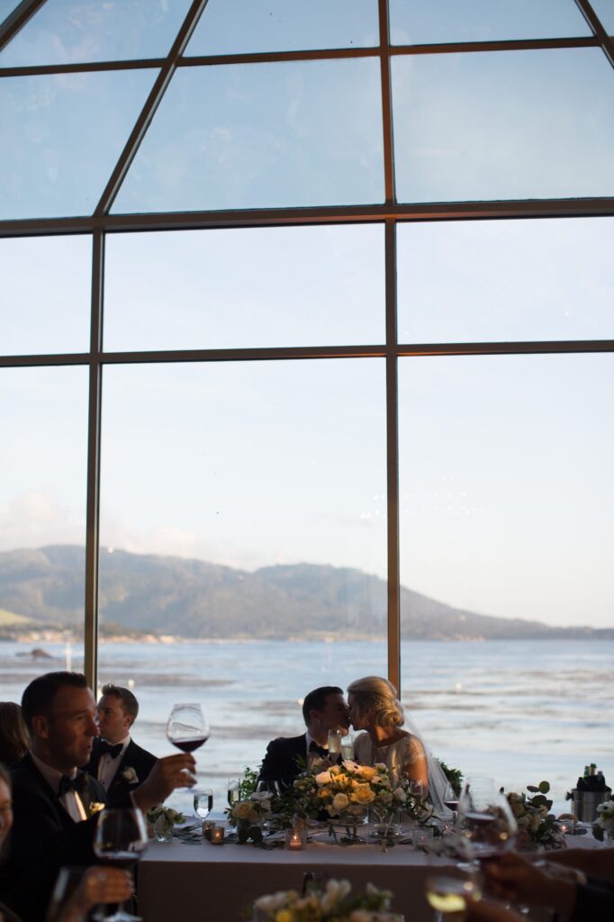 Husband and wife kiss at their reception dining in front of Pebble Beach with their guests seated at respective tables.