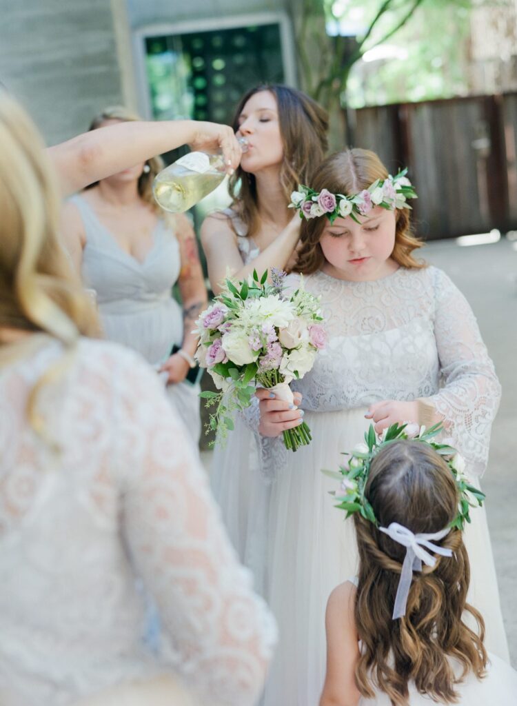 Bride drinks from a bottle while helping little girls at her wedding with their attires.