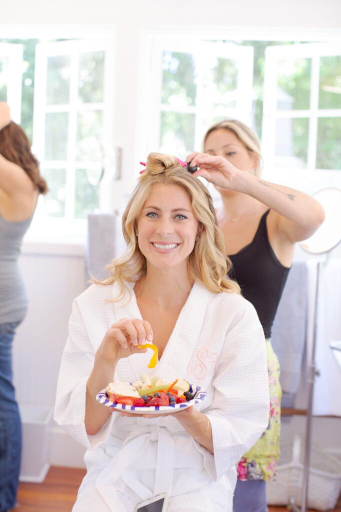 Bridesmaid adjusts the bride's hair as she eats a plate of fruits.