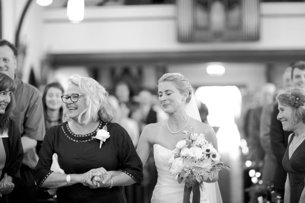 Bride holds a bouquet of flowers and smiles warmly while her mother smiles joyfully.