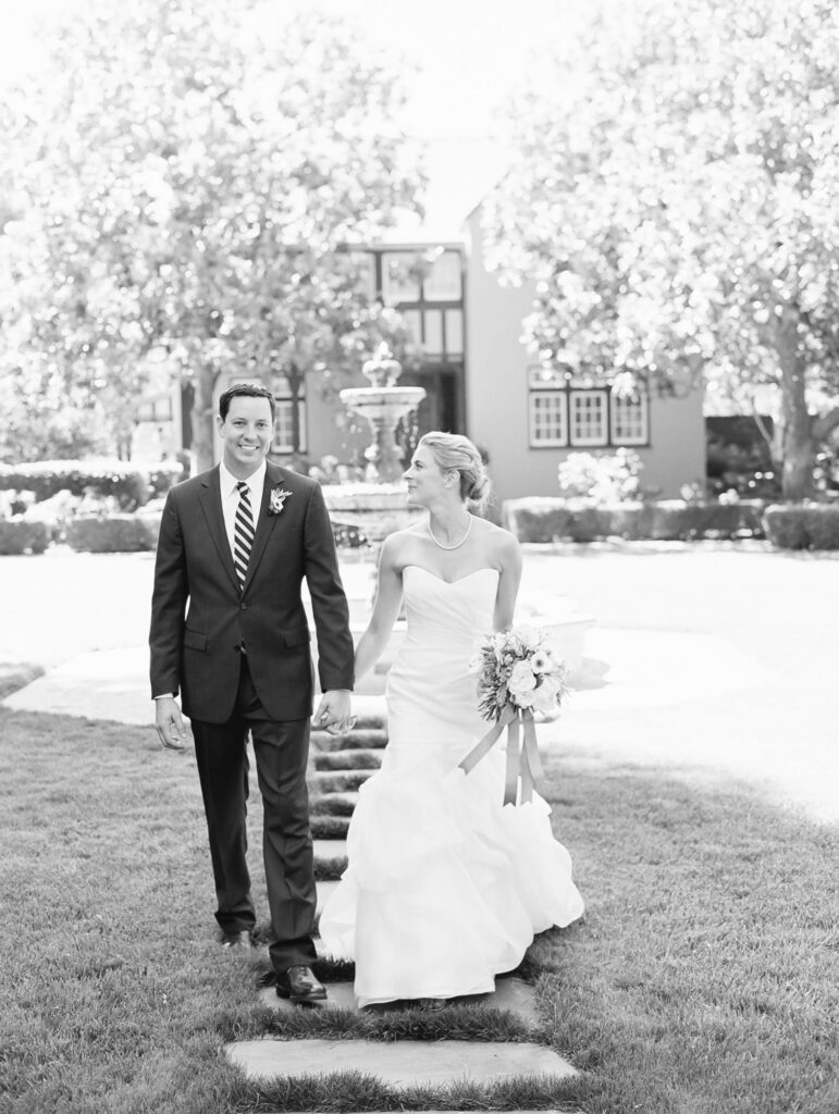 Newly married husband and wife holds hands in an outdoor garden or courtyard, with lush greenery and a beautiful fountain in the background.