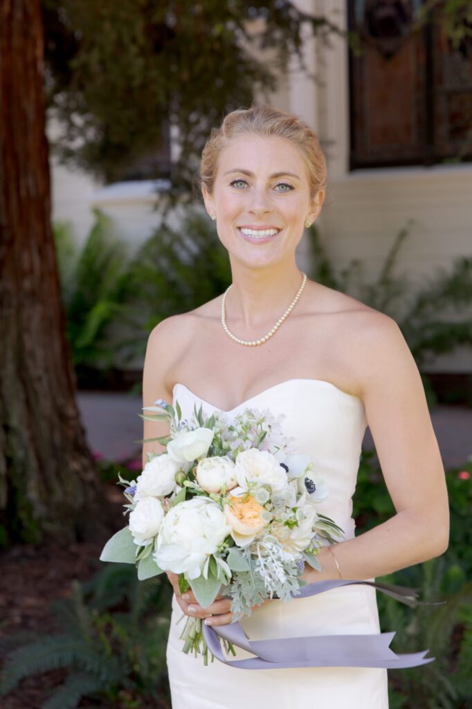 Elegantly dressed bride in wedding gown holds a bouquet of flowers and smiles at the camera.