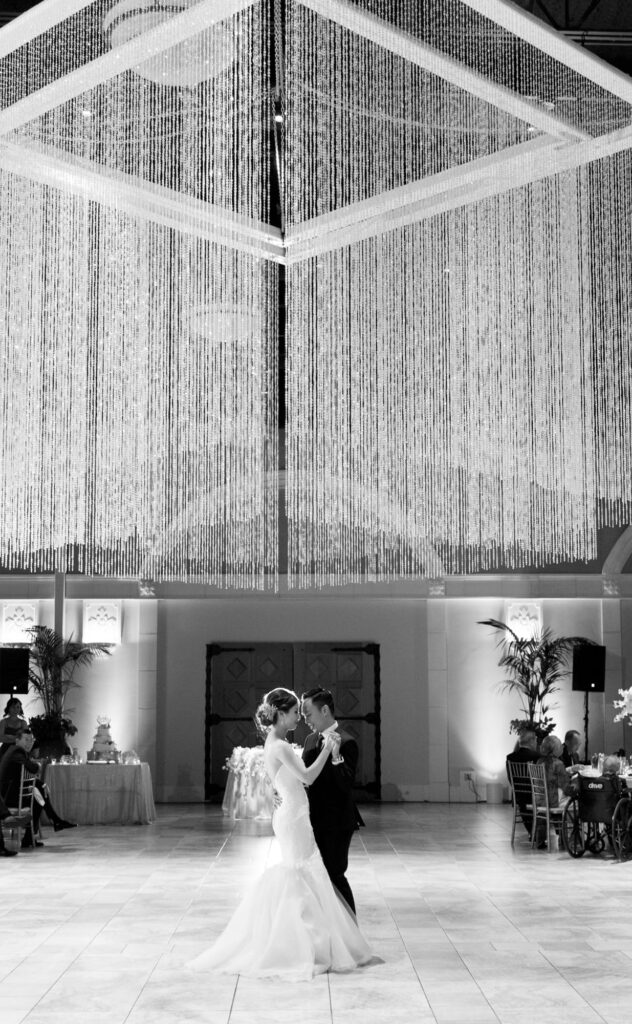 Bride and groom embrace each other for a dance below a giant elegant hanging thread-like light fixture.