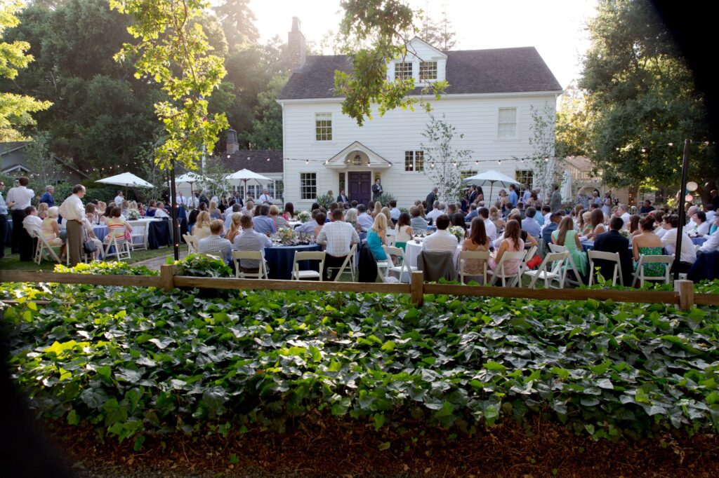 A vibrant, full-color image of an outdoor wedding reception held in the spacious, well-manicured grounds of a charming white estate.