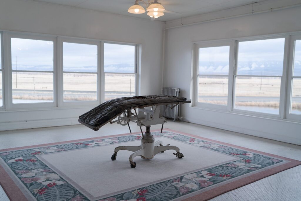 An old, white, mechanical examination or operating table with a black cushion on top, placed on a floral-patterned rug. 