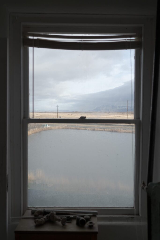 A window with a partially raised blind, looking out over a calm, reflective body of water and an expansive flat landscape. 