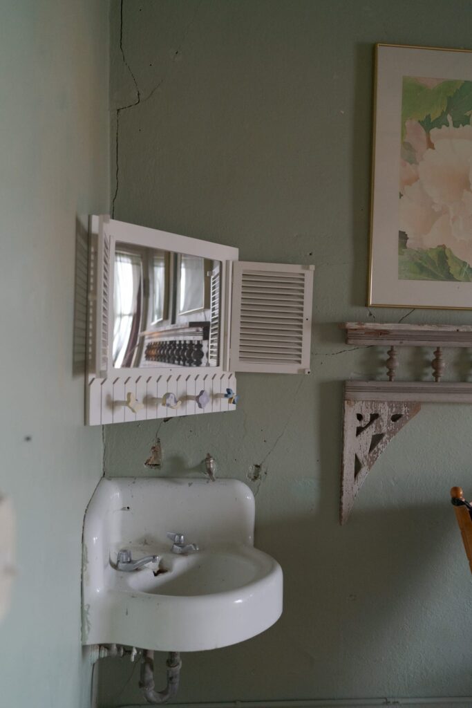 Close-up of a small, old sink mounted on a wall. Above the sink, there is a white medicine cabinet with small shutters and a mirror reflecting part of the room. 