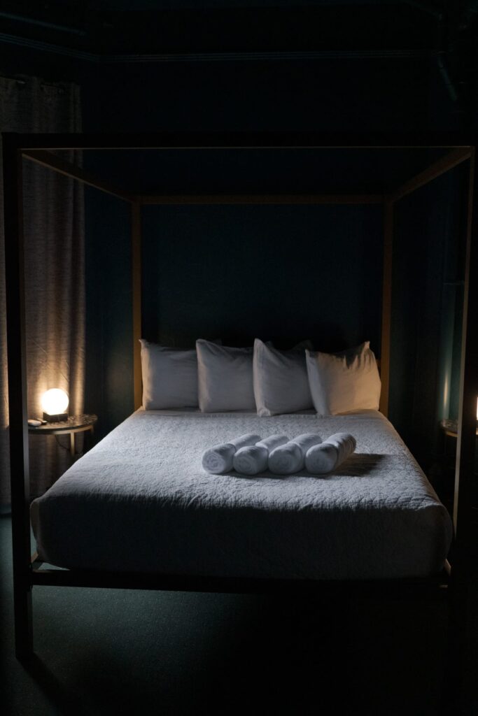 A dimly lit bedroom with a four-poster bed against a dark wall. The bed is made with white linens and four white pillows. Rolled white towels are neatly placed on the bed. 