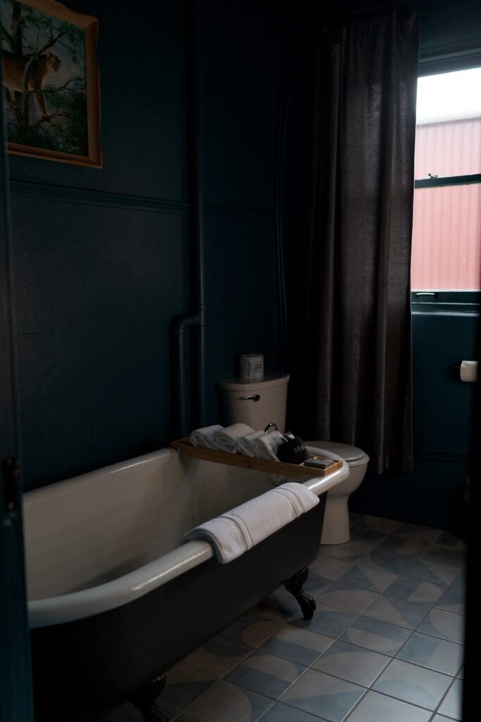 A bathroom with a dark blue clawfoot bathtub and a wooden bath tray holding white towels and a black toiletry bag