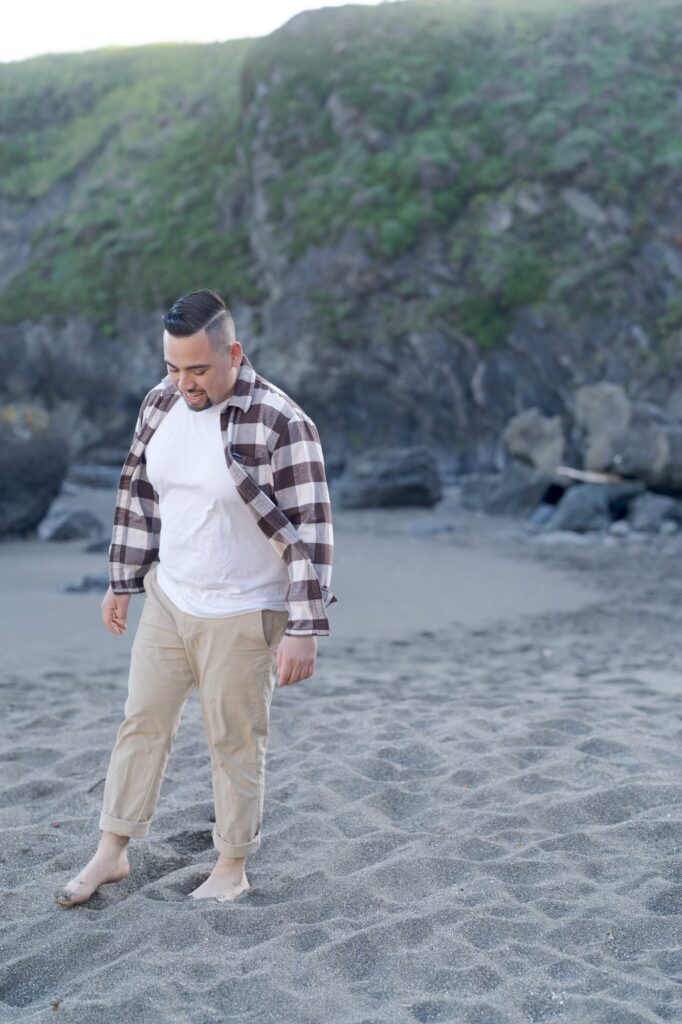 Man in white tshirt, khaki pants, and a brown checkered shirt walks on the beach.