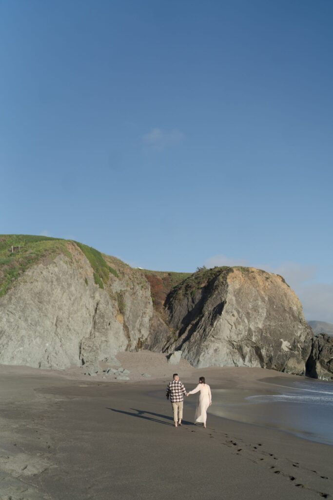 Newly engaged couple hold hands and walk in Bodega Bay while leaving footprints behind on the beach sand.