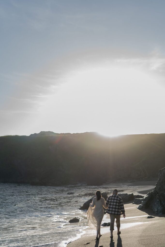 Newly engaged couple hold hands and walk towards the setting sun in Bodega Bay Beach.