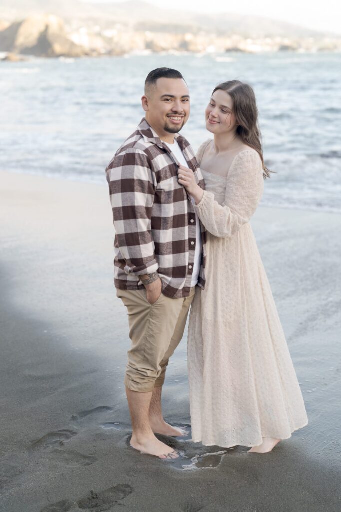 Man looks at the camera while his fiancee looks passionately at him at Bodega Bay Beach.