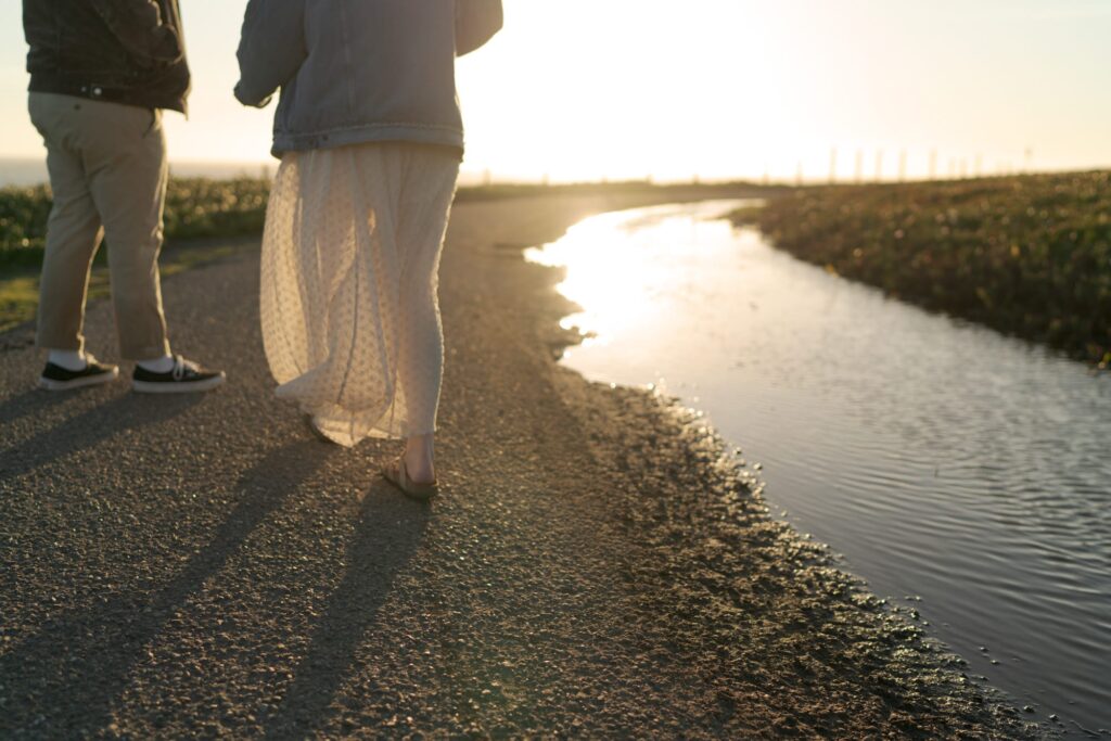 Man and woman in casual dress walk beside a shallow water stream during sunset.