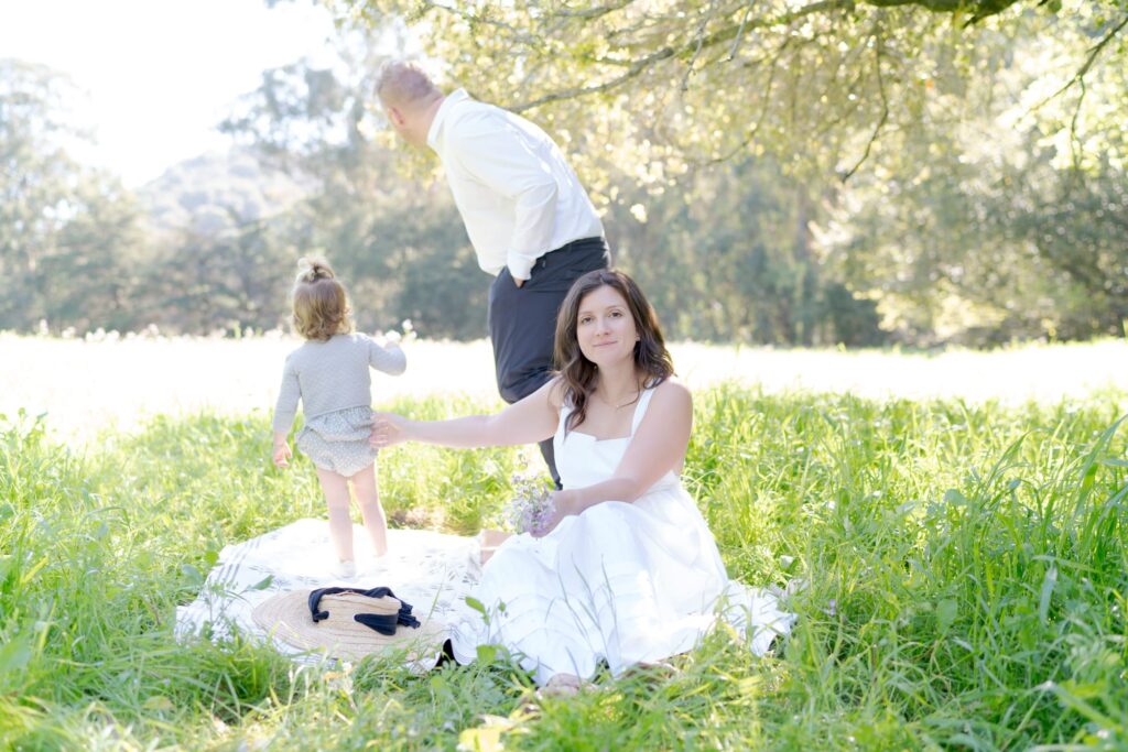 Jilian looks at the camera while her husband and daughter turn away and look at the grassy fields.