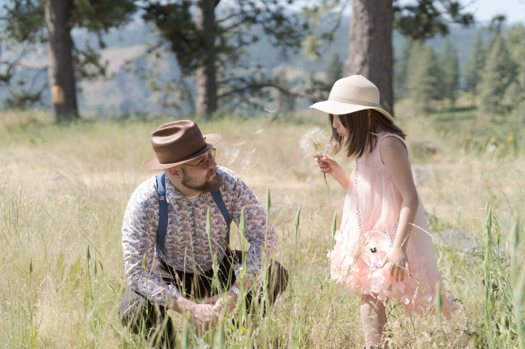 Dad watches her daughter blow into a blowball flower.