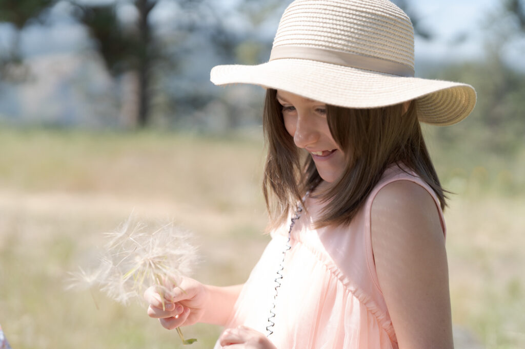 Girl in a cute pink dress and a sun hat holds a blowball flower.