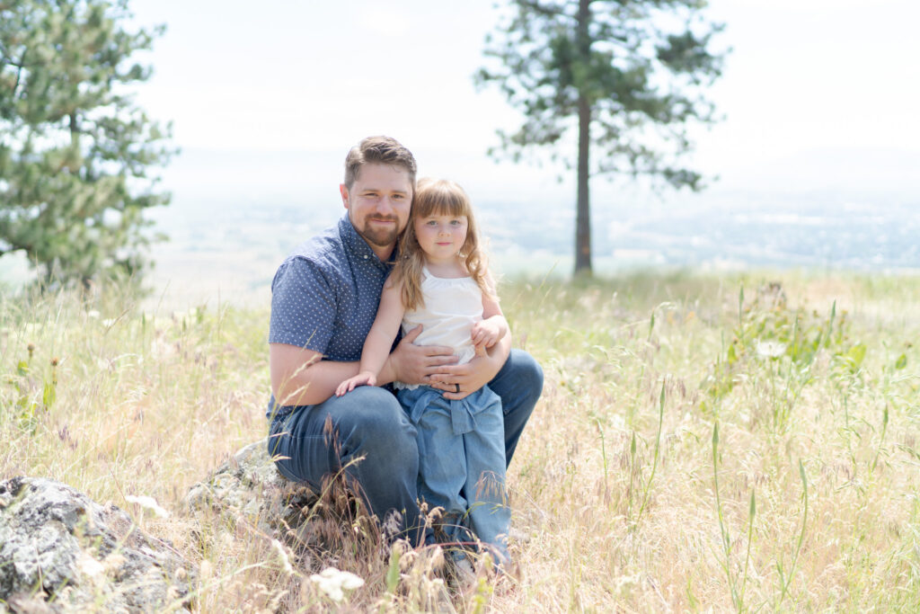 Dad lovingly holds his daughter amidst tall brown grasslands.