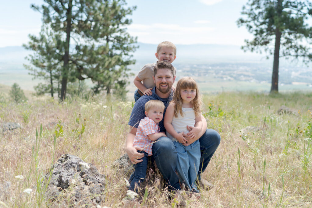 Dad poses for a picture with his children amidst tall grasses, wildflowers, and several pine trees,