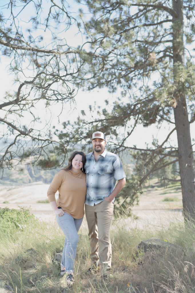 Husband and wife pose for a portrait in front of a big pine tree.