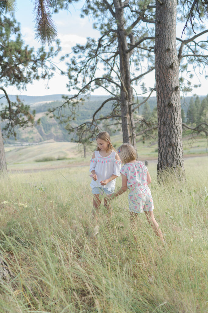 Little girls pluck flowers at a mountainous landscape under a partly cloudy sky.