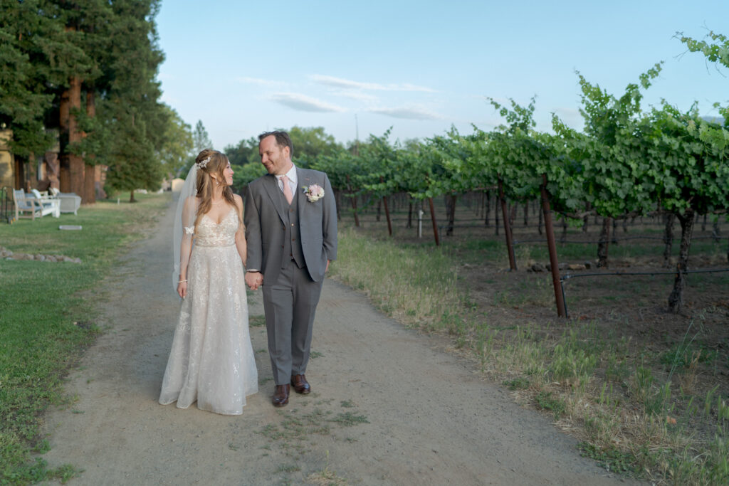 Bride and groom hold hands at the vineyard and look lovingly at each other.