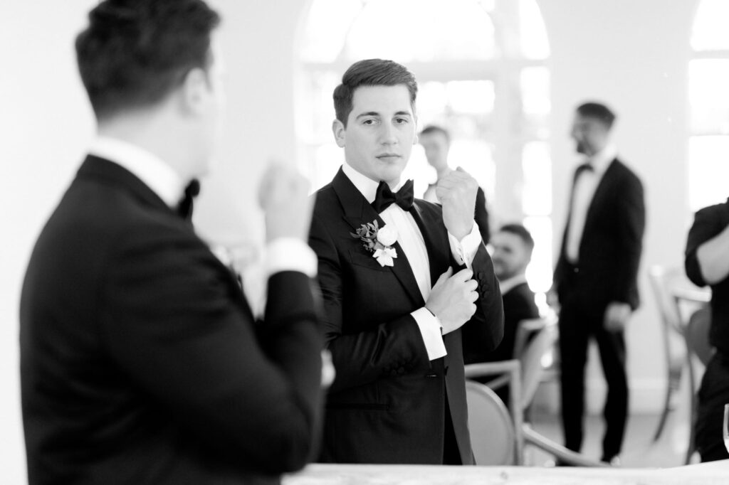 Handsome bridegroom in a black suit and bow tie looks at the mirror and adjusts his shirt.