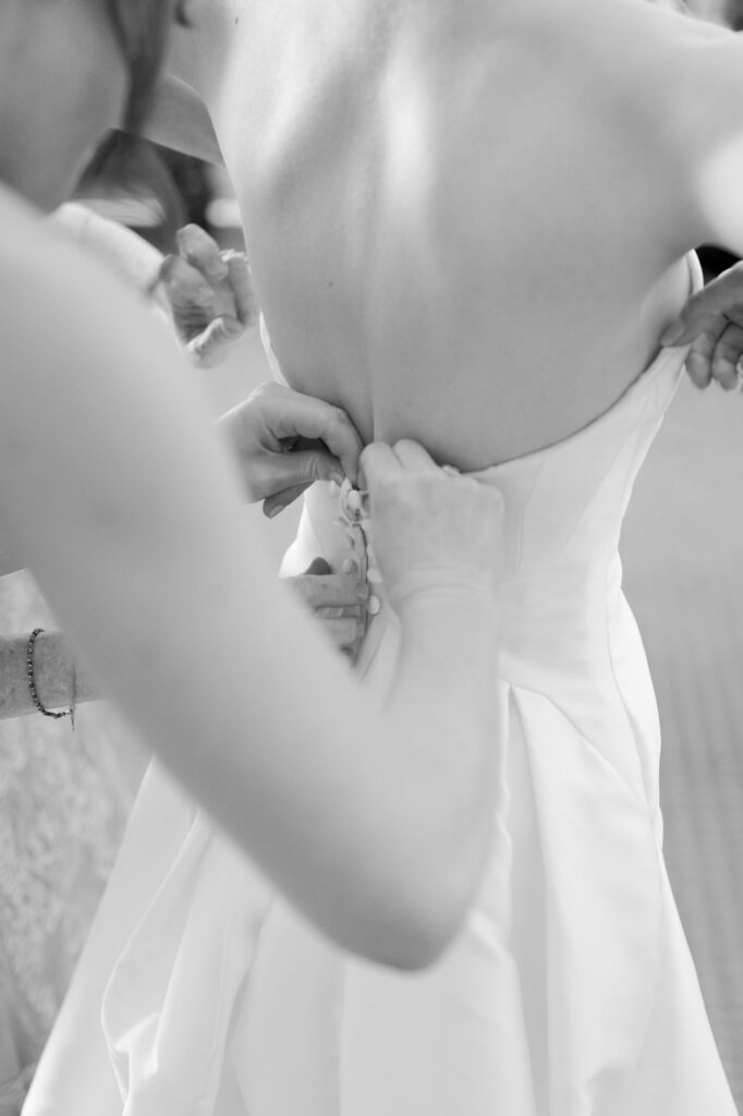 Bridesmaid helps the bride put on her white backless wedding gown.