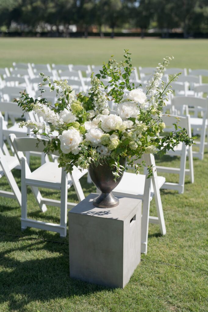 Beautiful white flower vase placed beside white chairs in an outdoor lawn.