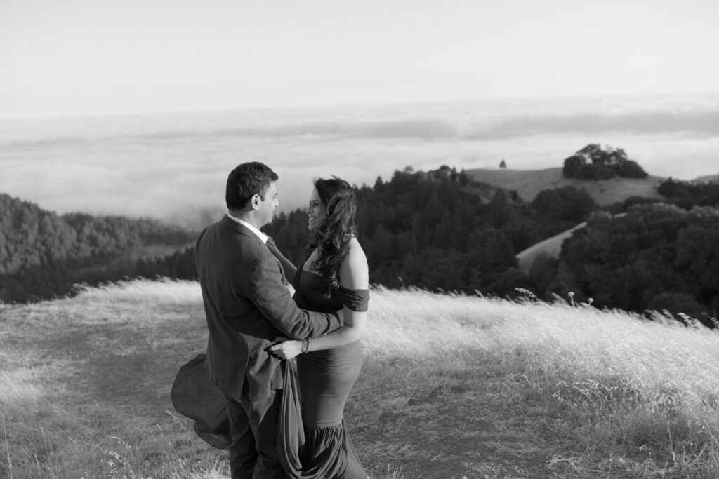 Husband and wife embrace each other at a mountain top above a picturesque sea of clouds in the background.