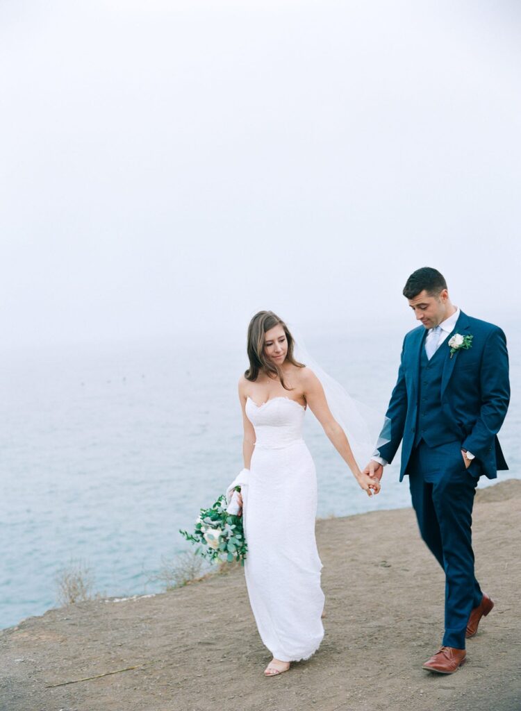 Newly married husband and wife hold hands and walk on the shores of Golden Gate Bridge.