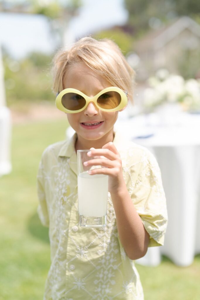 Blonde kid in a light-green shirt wears funky yellow glasses and grabs a glass of cold drink.