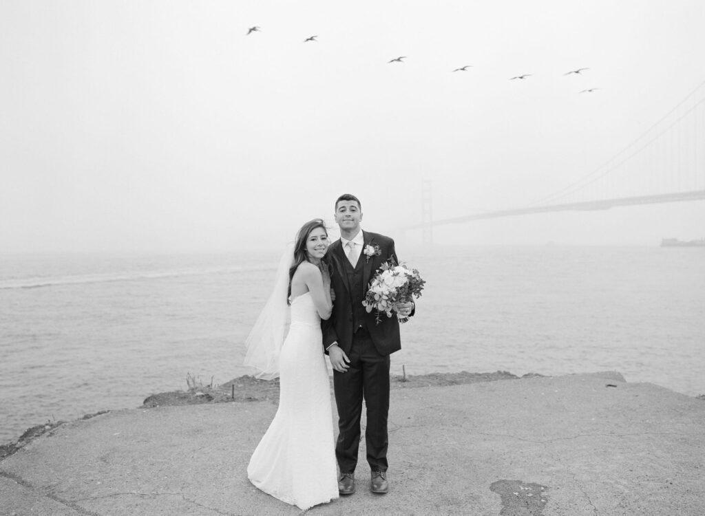 Tall bridegroom and his newly officiated wife pose in front of the Golden Gate Bridge in their elegant wedding attire.