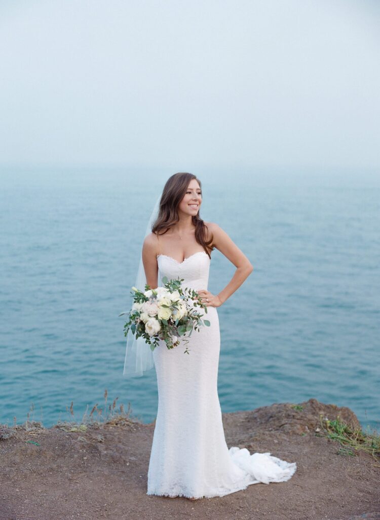 Newly married bride in her wedding gown holds a bouquet of white rises in front of a blue ocean background.