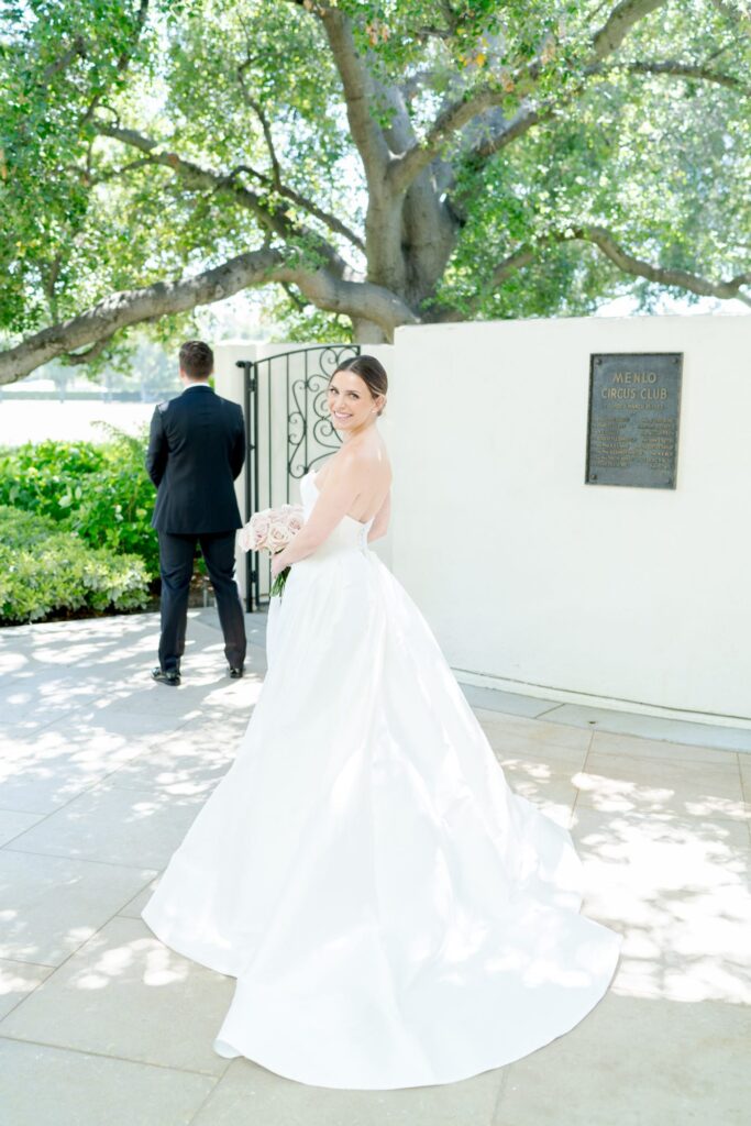 Bride in a sophisticated wedding gown turns and smiles at the camera with the groom in the background.