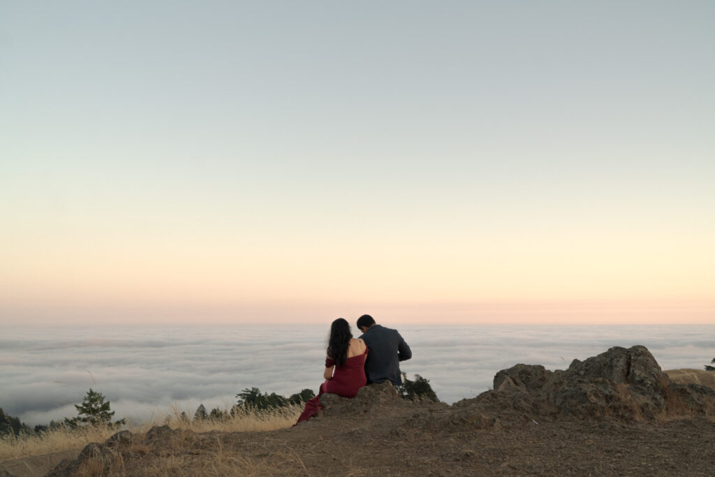 Husband and wife sit besides each other at a hilltop infront of an aesthetic background of floating clouds.